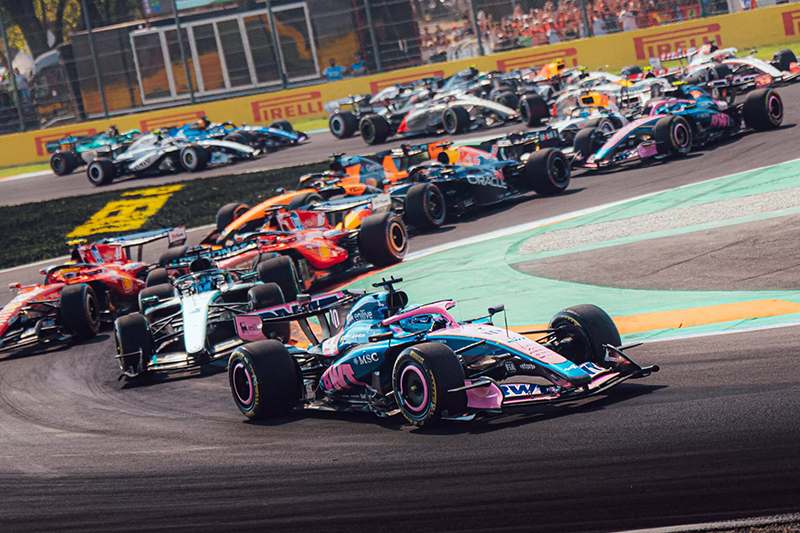 Scène de premier tour palpitante en Formule 1 : une vue de niveau de piste de plus d'une douzaine de voitures. Une voiture Alpine rose et bleue mène un groupe compact dans un virage à droite BWT Alpine