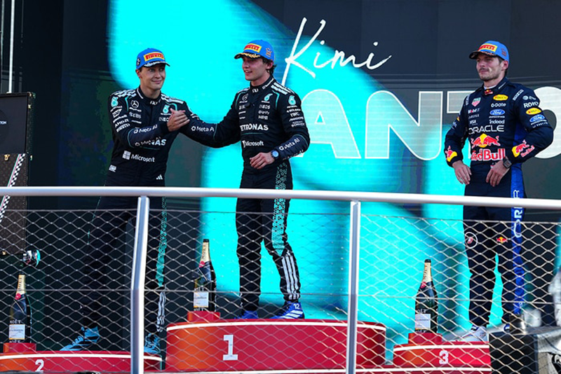 Podium F1 : Les pilotes célèbrent la victoire à côté d'un écran géant affichant 'Kimi AN'. Antonelli (Mercedes) serre la main d'un coéquipier, avec Verstappen (Red Bull) à droite. Andrea Kimi Antonelli